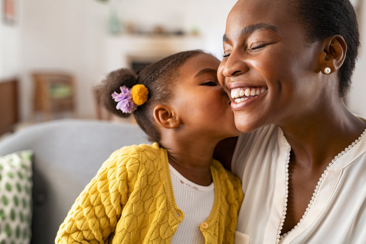 a woman of colour sits on the sofa with her little girl who is kissing her on her cheeck