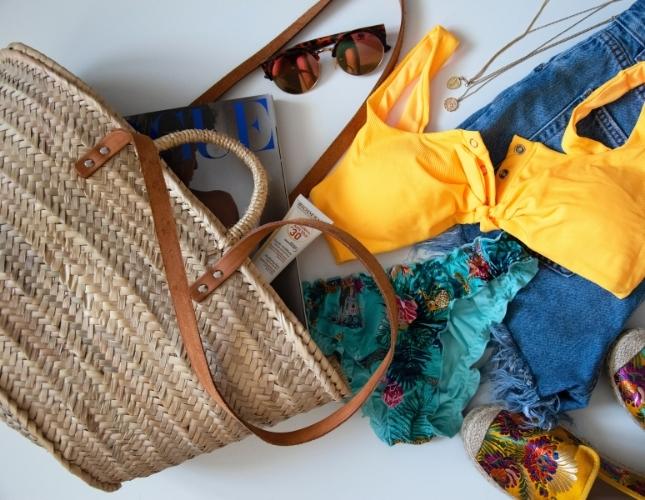 The contents of a straw beach bag - a bikini, sunglasses, and sunscreen - strown across a white background.